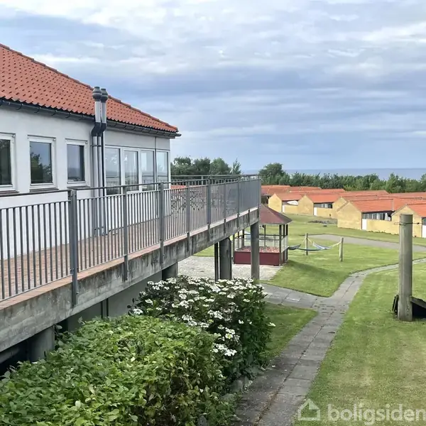 Et hus med rødt tag står på en forhøjning med balkon. I baggrunden ses flere ens huse og et åbent græsareal, mens himlen er overskyet.