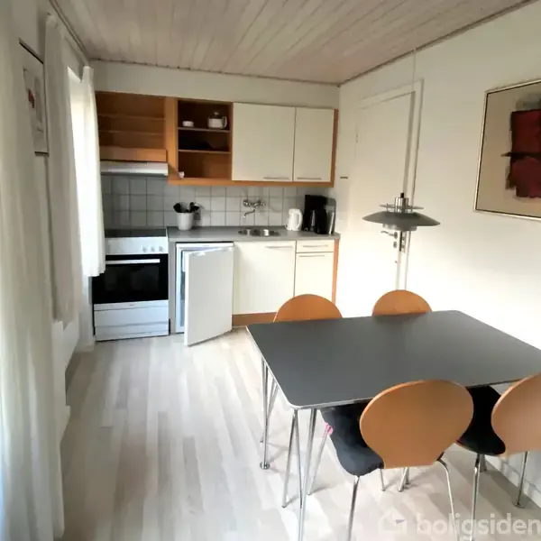 Et lille køkken med en grå bordplade og fire træstole. Et komfur og en håndvask er mod væggen. Rummet er lyst med gardiner og har en farverig kunstplakat på væggen.