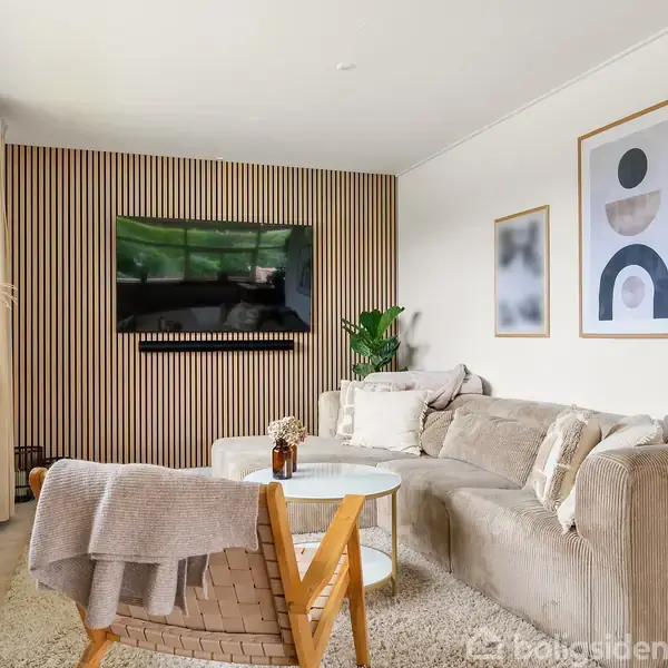 I en stue med lyse vægge og træbeklædte detaljer hænger et fladskærms-tv. En beige sofa med puder står ved et rundt sofabord. Dekorative billeder pryder væggen.
