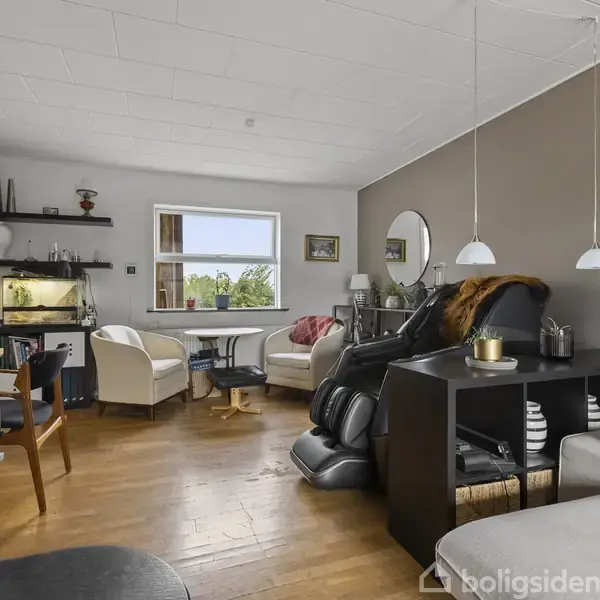 En hyggelig stue med en brændeovn til venstre, fisketank og hylde på bagvæggen, og moderne lænestole ved et stort vindue. Trægulve og neutrale vægfarver dominerer rummet.