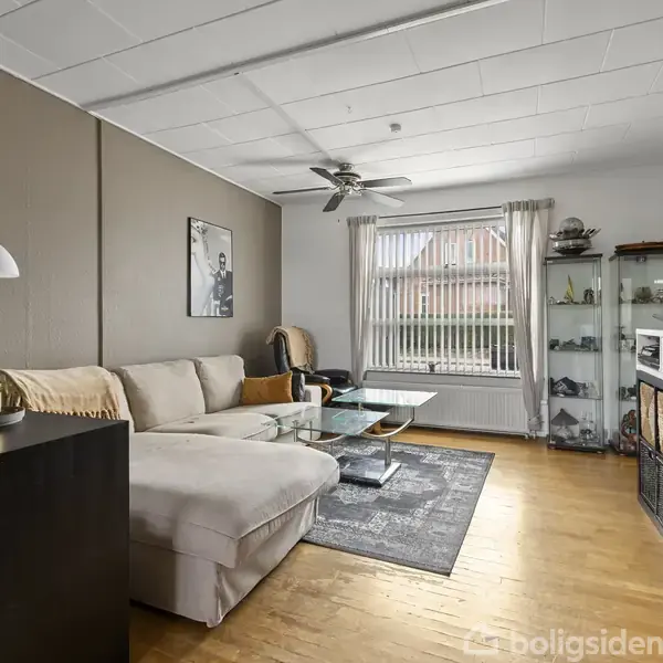 Sofa med lyse puder står i en stue, som har et sofabord og gulvtæppe. Der er hængende lamper, en reol og en stor fladskærm på væggen.