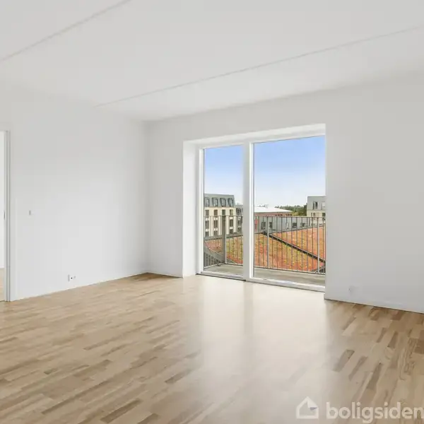 Et lyst værelse med trægulv står tomt. En skydedør fører til en balkon med udsigt over byens tage. Væggene er hvide og vinduet stort.