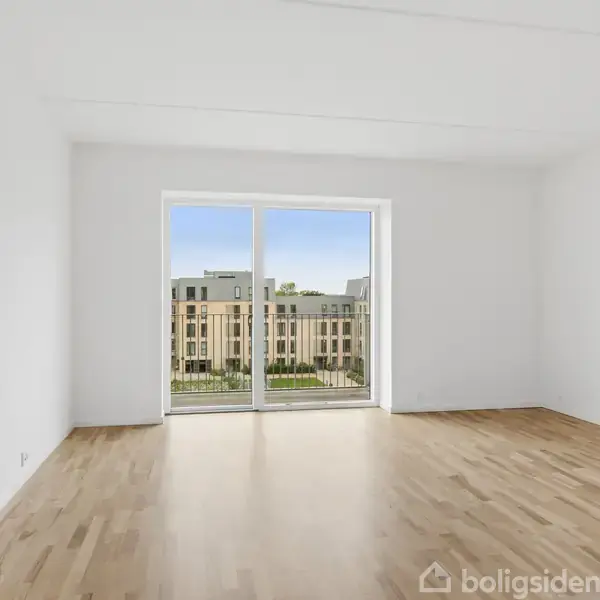 Et moderne køkken-alrum med spisebord og fire stole er placeret på et trægulv. I baggrunden ses et hvidt køkken, og til højre en grå sofa med gulvlampe, vase og sofabord.