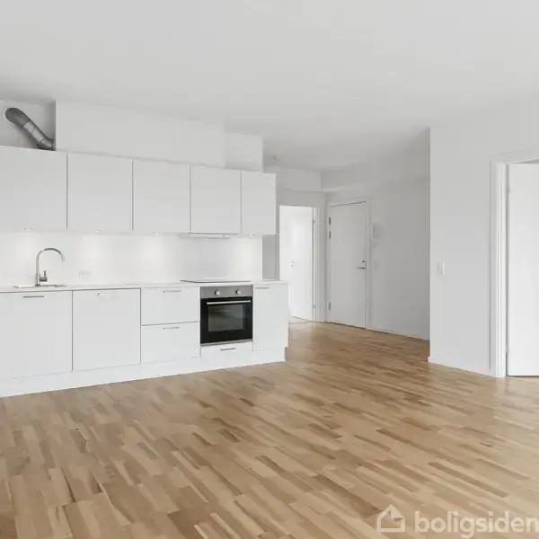 Stue med et rundt spisebord og stole, omgivet af et minimalistisk køkken på venstre side. En sofa med planter i baggrunden og en buet lænestol ved et stort vindue.