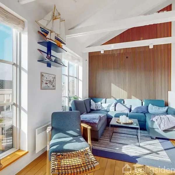 En blå sofa med puder står i et lyst rum med træpanelvæg. Ved siden af sofaen er et bord med lys. Et stort vindue lader sollys strømme ind, og der hænger en modelsejlbåd på væggen.