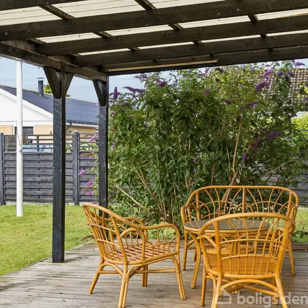 Et sæt bambus havemøbler står på en træterrasse under en pergola. Møblerne vender mod en stor grøn busk, som tilføjer privatliv til den indhegnede have.