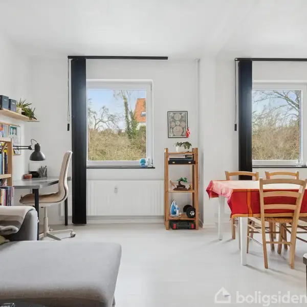 En stue med en grå sofa, et skrivebord med stol og en reol fyldt med bøger. To vinduer med udsigt til buske og et spisebord med rødt dug.