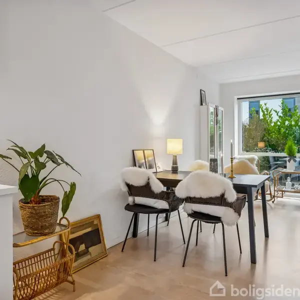 Stue med spisebord, hvor stole er dækket med fåreskind. En åben dør fører til en altan med planter. Indretningen inkluderer en kaffemaskine og kunst på gulvet.