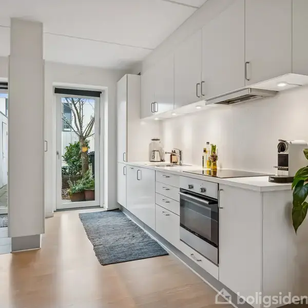 Et moderne køkken med hvide skabe har en ovn, kogeplade og køkkenmaskiner. Det åbner ud mod en terrasse med grønne planter, omgivet af store glasdøre, der slipper lys ind.