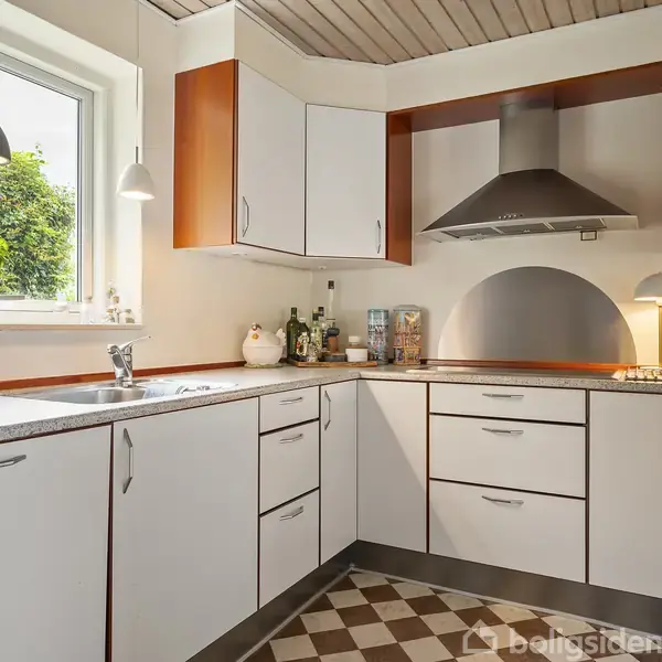 Et hjørnekøkken med hvide skabe og stålgreb. En emhætte hænger over komfuret. På bordet står en lampe og krydderier. Et vindue med plante giver udsigt til grønne træer.