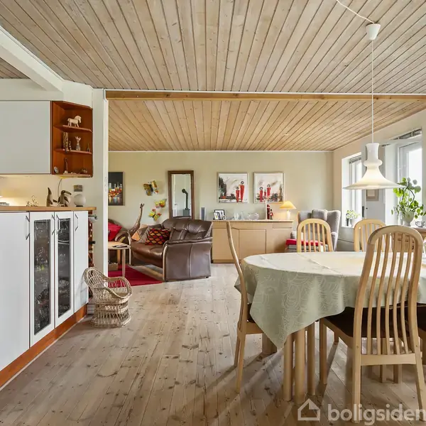 Spisebord med dug står i et rum med trægulv og træloft; omkranset af lyse træstole. I baggrunden ses sofa, reoler, billeder og lamper, der skaber en hyggelig atmosfære.