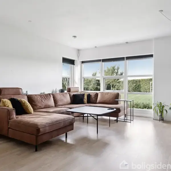 Brun lædersofa står i en lys stue med store vinduer. Et sofabord er placeret foran sofaen. Rummet har også planter, minimalistisk dekoration og en moderne lampe i loftet.