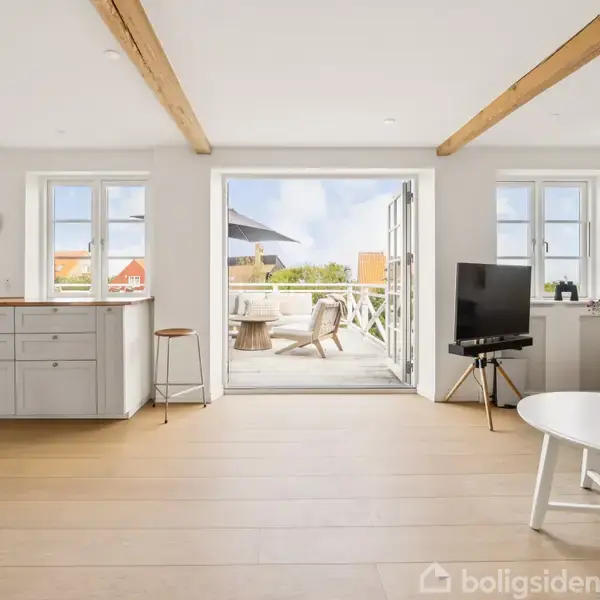 Stue med sofa og tv står foran åbne døre til en terrasse med udendørsmøbler. Lyse trægulve, synlige loftsbjælker og et moderne, minimalistisk design præger rummet.