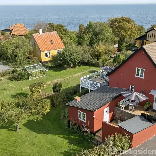 Rødt hus med spidst tag står stille i en frodig have, der omfatter en terrasse og havegang. I baggrunden ses flere huse og en udsigt over havet.