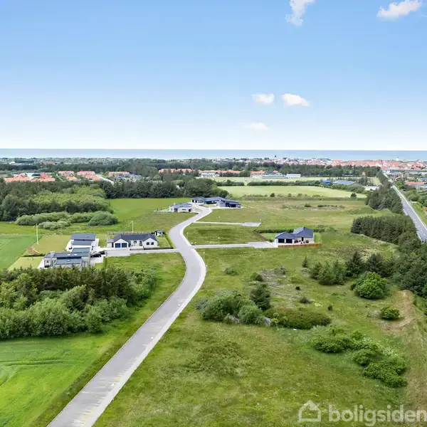 En landevej strækker sig gennem et åbent grønt landskab med spredte huse. I horisonten ses en by med tætliggende bygninger, omgivet af marker og træer under en klar blå himmel.