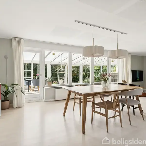 Spisebord med stole står midt i en lys stue. Rummet er omgivet af store glaspartier, planter og et hyggeligt sofahjørne med vægmonteret TV.
