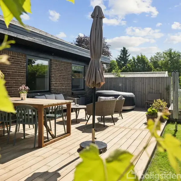En træterrasse med et spisebord, stole og en parasol i en have ved et murstenshus. En sofa og en jacuzzi ses i baggrunden med grønne planter omkring.