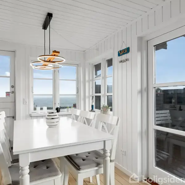 Et hvidt spisebord med seks stole står i et lyst rum. Moderne lamper hænger over bordet. Rummet har trægulv, hvide vægge og store vinduer, der giver udsigt til havet.