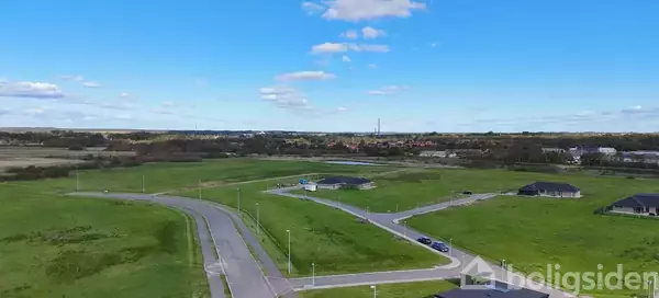 Grønne marker strækker sig under en klar blå himmel, mens en snoet asfalteret vej fører gennem landskabet. Enkelte huse ses spredt i det fjerne.