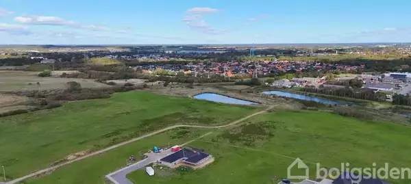 Et græsområde med en igangværende byggeplads i forgrunden; en vej fører til en lille by med huse og træer i baggrunden.