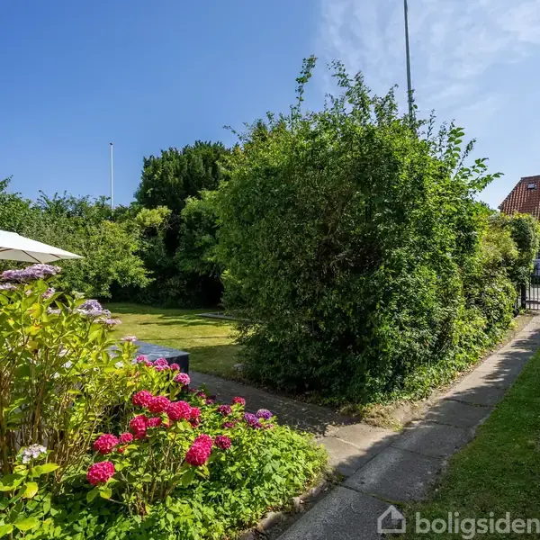 En velplejet have med blomstrende buske og en grussti. En hvid parasol står i græsset med en hvid bygning til venstre og et hus med rødt tag i baggrunden.