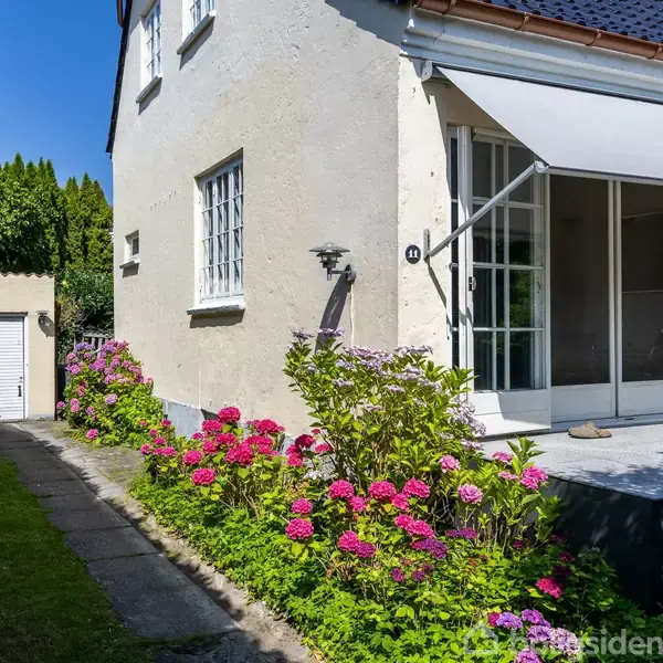 Hus med hvide vægge og en markise over terrassen. Blomstrende pink hortensiaer langs husets fundament. Stenbelagt gangsti fører til en hvid garageport omgivet af hæk.