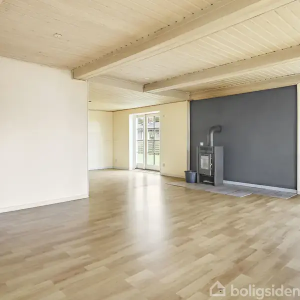 Et lyst og rummeligt rum med trægulv. En brændeovn står mod en grå væg. Vinduer og dør til venstre giver udsigt til udendørsområdet. En åbning fører til badeværelse.