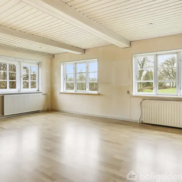 Et lyst rum med trægulv og hvide lofter viser store vinduer, der oversvømmer rummet med naturligt lys. Radiatorer er placeret under vinduerne mod udsigt til en grøn have.