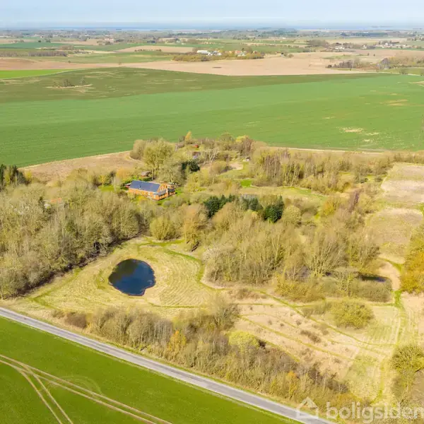 En gård ligger omgivet af grønne marker og en lille sø, set fra luften. Veje strækker sig langs markerne, og landskabet er åbent og landligt.