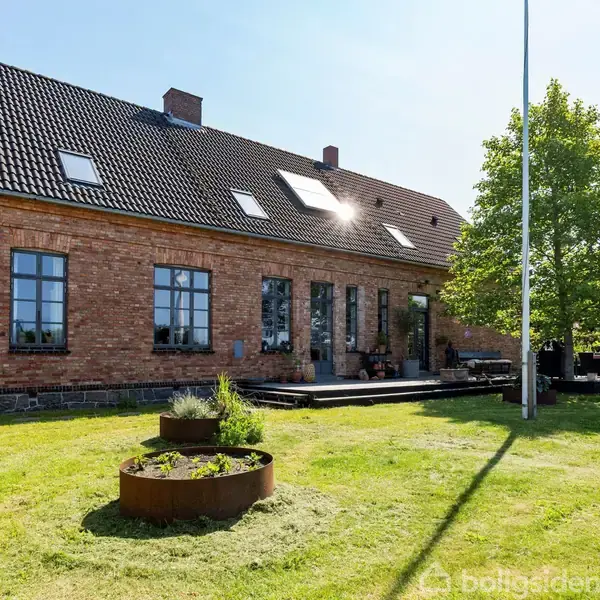 En rødstensbygning med sort tegltag står omgivet af en velplejet græsplæne og et træ. Foran huset er der runde metalbed med planter.