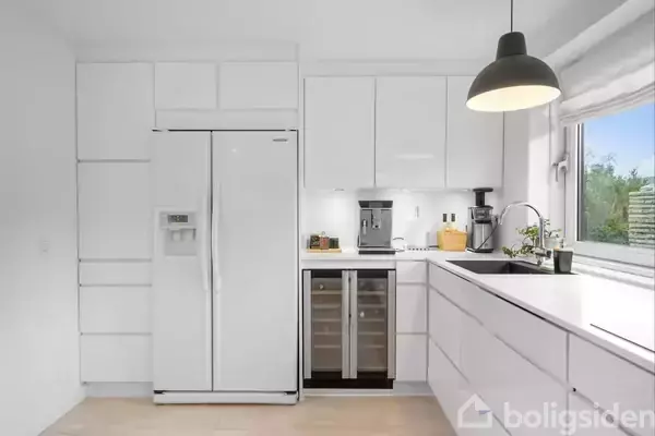 Køleskab står i et moderne, hvidt køkken med strømlinet design, ved siden af vinreol og kaffemaskine, under et stort vindue der giver udsigt til grønne træer udenfor.