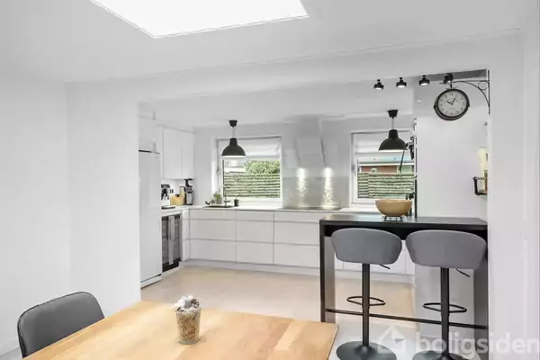 Et moderne køkken med hvide skabe og stålarmatur. To grå barstole står ved en bardisk. Et spisebord befinder sig i forgrunden, og et ur hænger på væggen.