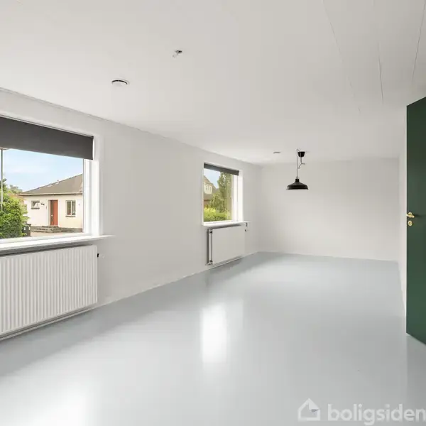 Et lyst rum med hvidt gulv og vægge. To store vinduer med udsigt til tage og træer giver naturligt lys. En sort lampe hænger fra loftet.