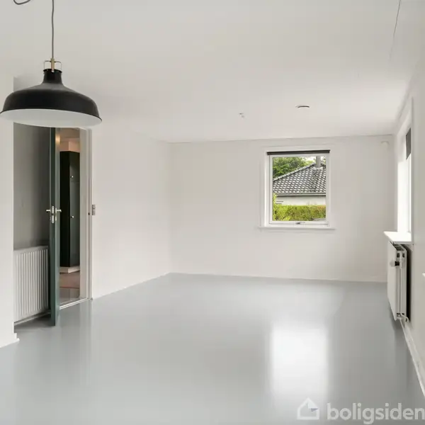 Et tomt rum har en hængende sort lampe, med et åbent dør ind til en anden sektion af huset. Gennem vinduerne ses et udsigt til en have med træer.