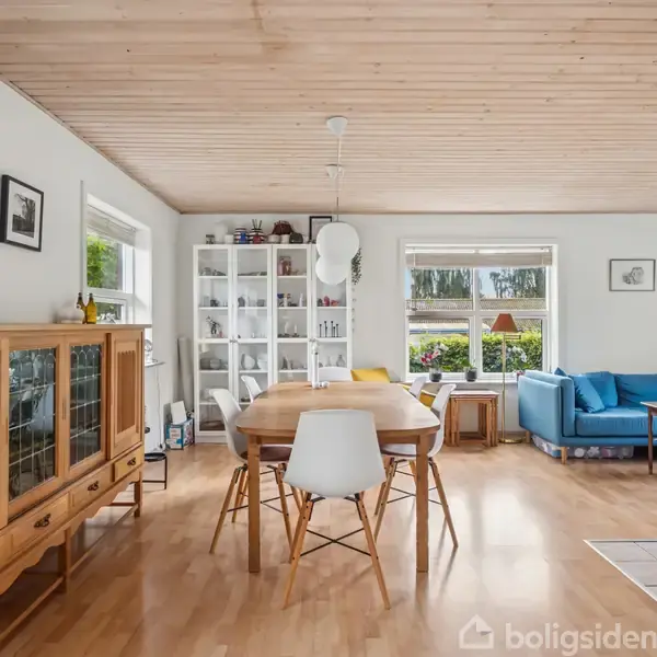 Et spisebord står midt i en lys stue. Rundt det er der stole, en reol fuld af genstande, et skab, og en blå sofa ved vinduet.