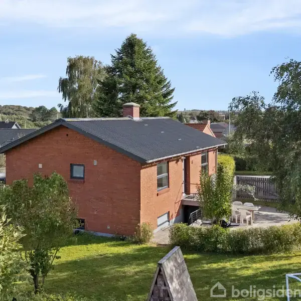 Rødt murstenshus står stille i en frodig have med græs og træer, omgivet af boligområde. Terrasse med havemøbler tæt på husets indgang. Ingen synlig tekst i billedet.