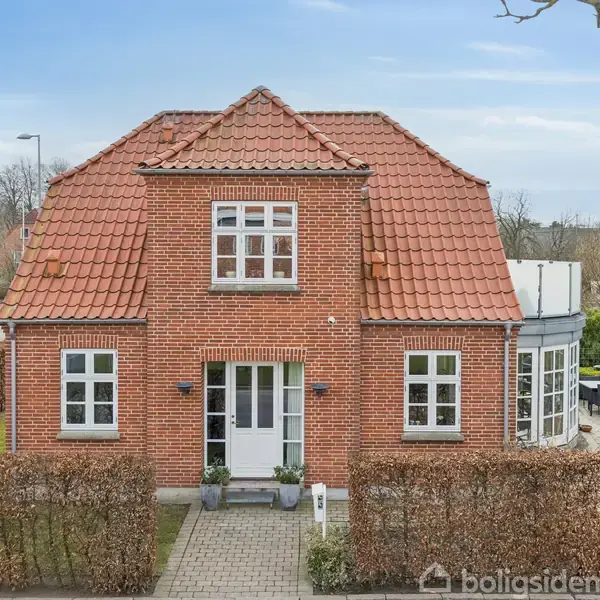 Rødt murstenshus med rødt tegltag, står stilfuldt. Hækken omkranser indkørslen, der leder til hoveddøren. Villaen har flere vinduer og en tilbygning til højre i billedet.