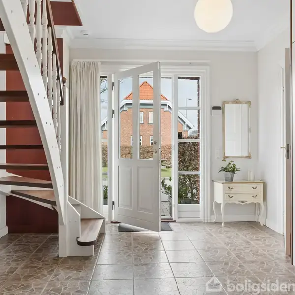 Trappe med trætrin fører opad i en lys entré. Terrassedør står åben mod have med rødt hus i baggrunden. Skab og lille kommode står ved siden af døren.