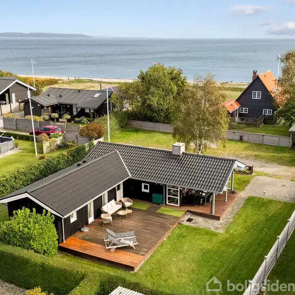 Et sommerhus med fladt tag og en træterrasse står i en have nær havet. Omgivet af andre mindre huse og haveanlæg.