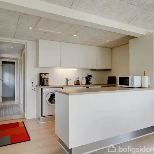 Køkken med en hvid køkkenbordplade, vaskemaskine under bordet, kaffemaskine, brødrister, og mikroovn. Rummet har træpanelvægge og et farverigt gulvtæppe nær en lang gang.