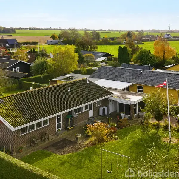 Et parcelhus med græsplæne og flagstang i en forhave. Omkringliggende huse og marker i en landsbylignende setting.