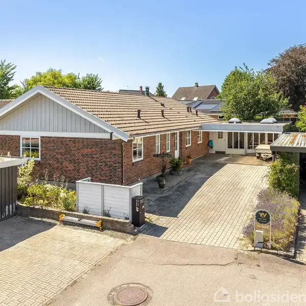 En énetages murstensvilla med garage og carport, omkranset af blomsterbede. Beliggende i et villakvarter med grønne træer. Der holder en bil parkeret under carporten ved siden af villaen.