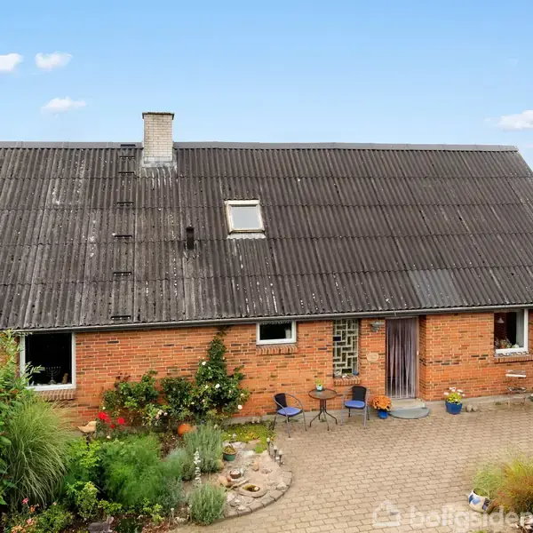 Et gammelt, rødt murstenshus med en sort, slidt tag, står stille i en lille gårdhave med blomsterbede og havemøbler. Baggrund præges af marker og en blå himmel.