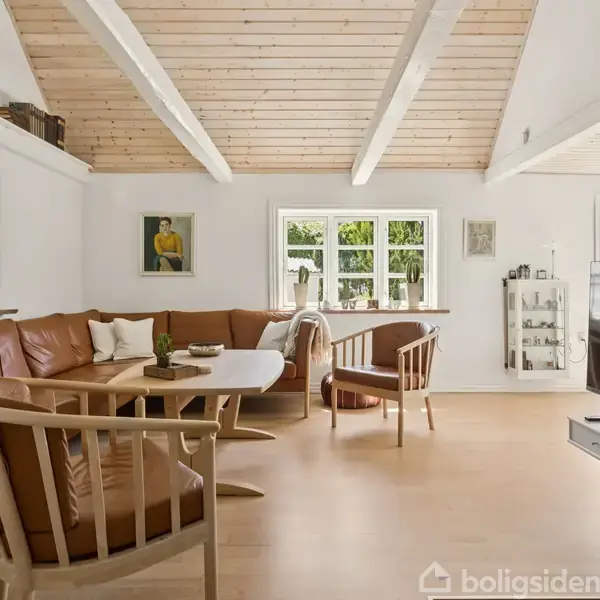 Brune lædermøbler står i en lys stue med trægulv og loftsbjælker. Til venstre ses en lædersofa og stole omkring et bord. Rummet har vinduer, kunst på væggene og et TV.