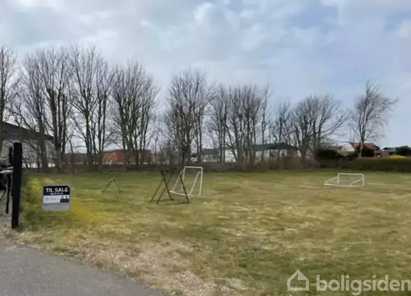 Græsfelt med fire små fodboldmål står stille; omgivet af træer og nogle få bygninger. Et skilt viser teksten: "TIL SALG" sammen med Boligsidens telefonnummer.