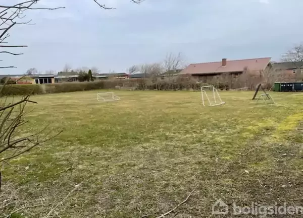 Fodboldmål står på græsplæne i en åben have. Omgivet af buske og træer, i et villakvarter med huse i baggrunden.
