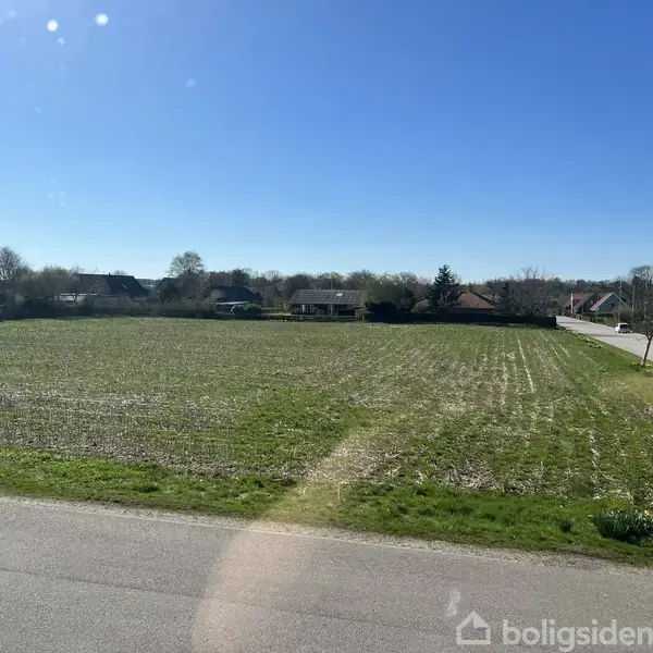 En grøn mark med spredt vegetation ligger roligt ved siden af en vej. I baggrunden ses lavtliggende huse omgivet af træer under en klar blå himmel.