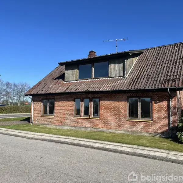 Murstenshus står ved en vej, med græsplæne omkring. Stort skråt tag, vinduer på både stueetage og førstesal. Blå himmel, træer i baggrunden, boligområde.