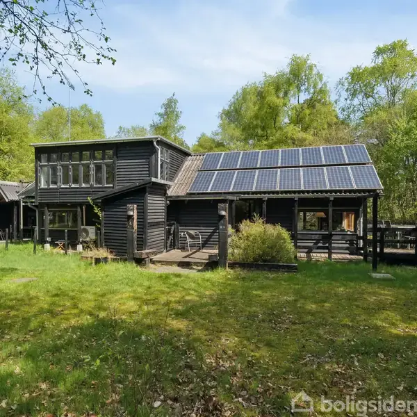 Et sort træhus står omgivet af grønne træer med solpaneler på taget i en frodig have.