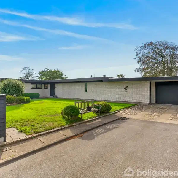 Et moderne, lavt hus med fladt tag står på en velplejet græsplæne. En bænk og buske er foran, og en garage er på højre side, omgivet af et stilrent haveanlæg.